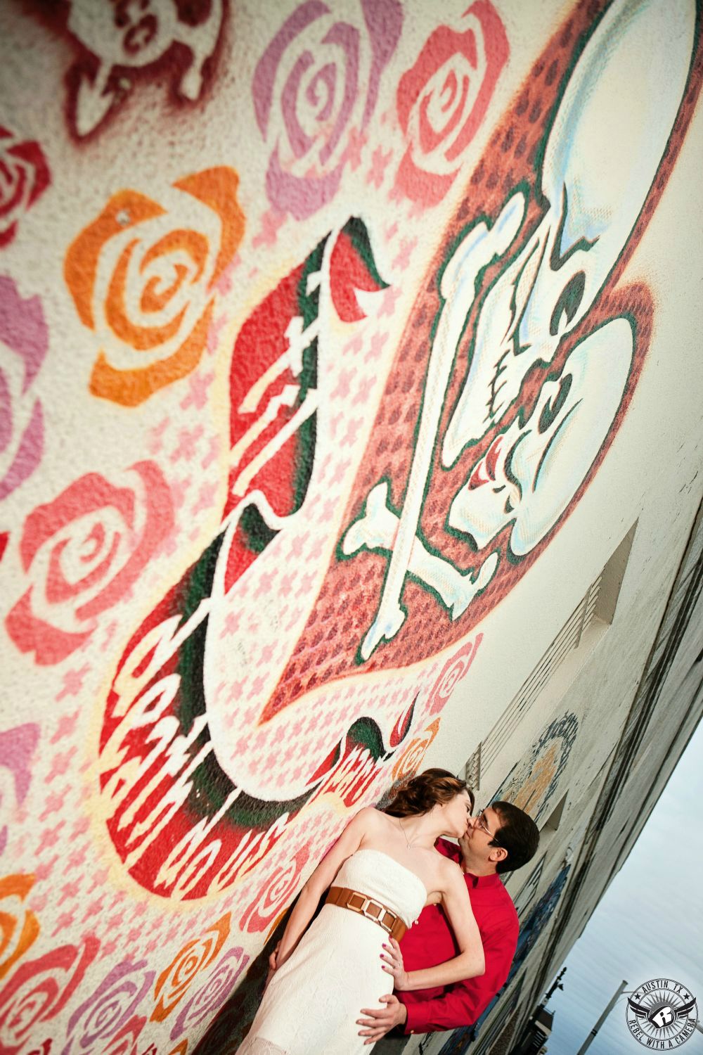 til death do us part mural with two skulls and roses with engaged couple leaning on wall wearing a white dress with a tan belt and a red button up shirt kissing in downtown austin